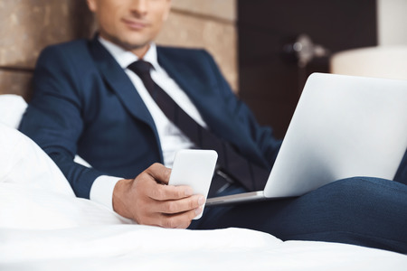 Businessman in formal suit using his phone and laptop while sitting on bedの写真素材