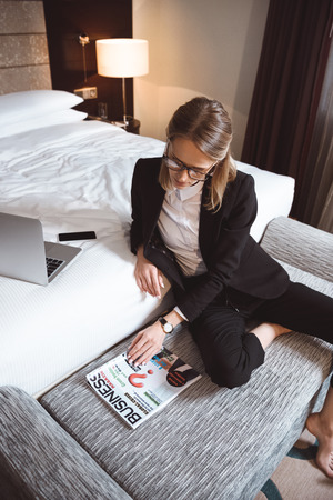 high angle view of young businesswoman in eyeglasses reading magazine in hotel roomの写真素材