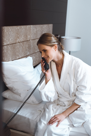 smiling young woman in bathrobe talking by phone in hotel roomの写真素材