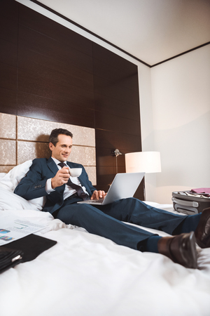 Young businessman in formal suit using his laptop and holding cup of coffee, while sitting on bed in hotel roomの写真素材