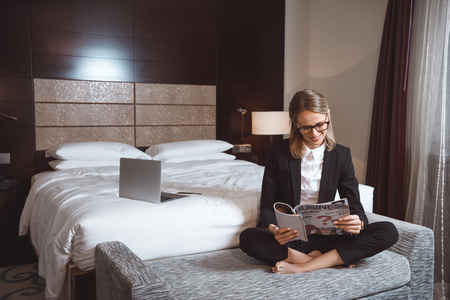 smiling young businesswoman in eyeglasses reading magazine while sitting in hotel roomの写真素材