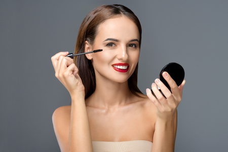 gorgeous young woman applying makeup and smiling at camera isolated on greyの写真素材