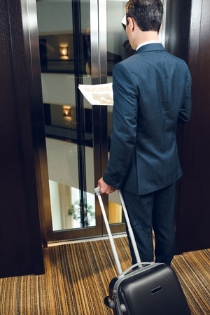 businessman in hotel corridor with a suitcase, waiting for elevator and reading newspaperの写真素材