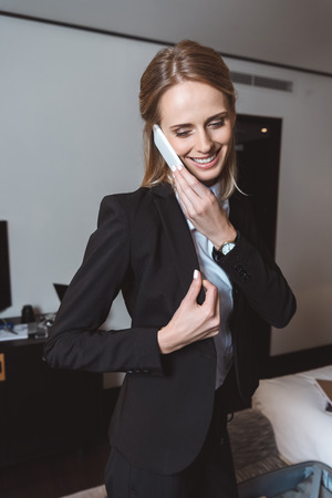 smiling young businesswoman talking on smartphone while wearing suit jacket in hotel roomの写真素材