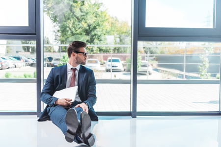 young businessman in suit and eyeglasses holding laptop while sitting on floor in officeの写真素材