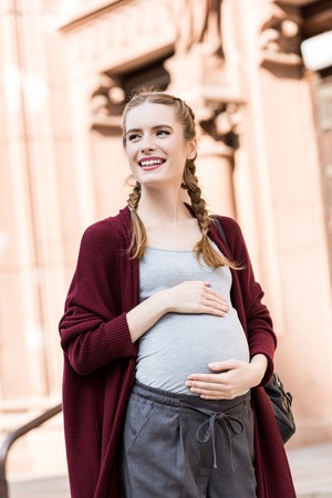 portrait of happy pregnant woman touching belly and looking away on streetの写真素材