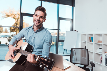 handsome smiling young businessman playing guitar in officeの写真素材