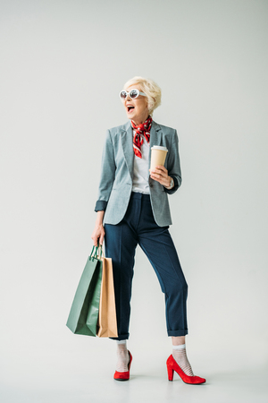happy senior woman in jacket and sunglasses with shopping bags and coffee, isolated on greyの写真素材