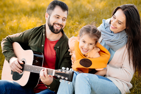 Family with daughter relaxing on a grassy hill while father playing the guitarの写真素材
