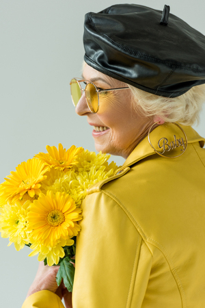 stylish senior woman in yellow jacket with bouquet of yellow flowers, isolated on greyの写真素材