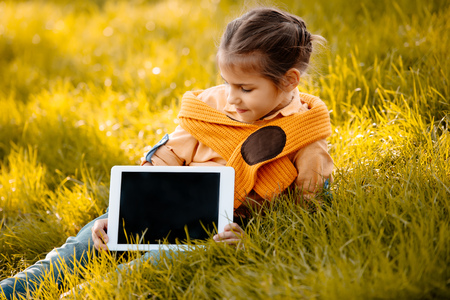 Little child sitting on autumn grass and showing digital tablet screenの写真素材