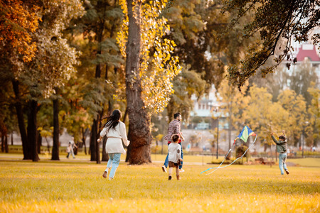 Family with two children flying a kite and running across grassy lawn in autumn park