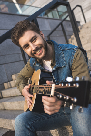 portrait of smiling man looking at camera and playing guitar while sitting on stairs on streetの写真素材