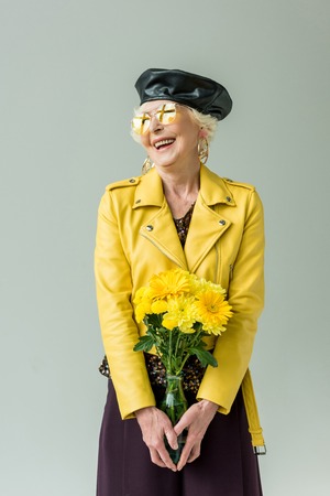 stylish senior woman in yellow leather jacket with bouquet of yellow flowers, isolated on greyの写真素材