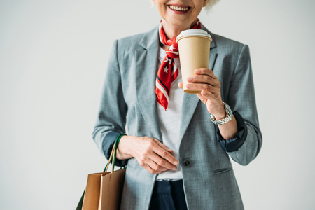 cropped view of senior woman in jacket and sunglasses with shopping bags and coffee, isolated on greyの写真素材