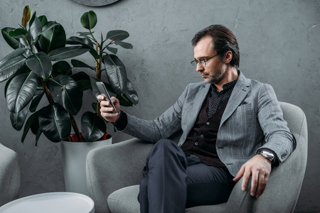 serious businessman in eyeglasses using smartphone while sitting on armchairの写真素材
