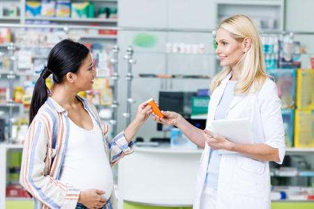 smiling pharmacist with digital tablet giving pills to pregnant woman in drugstoreの写真素材