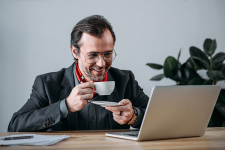 smiling businessman in eyeglasses drinking coffee and using laptop at workplaceの写真素材