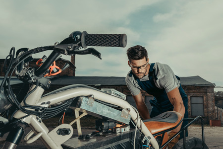 repairman in goggles repairing vintage motorcycle standing outside at garage