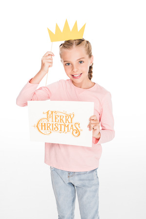 Cheerful child playing with cardboard crown and holding a card with Merry Christmas text, isolated on whiteの写真素材