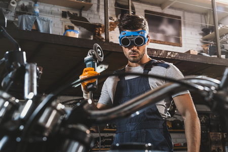 young handsome mechanic in goggles with circular saw in workshop with motorcycleの写真素材