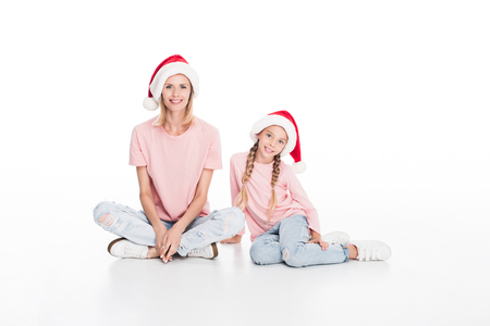 Happy mother and daughter in santa hats sitting on floor, isolated on whiteの写真素材
