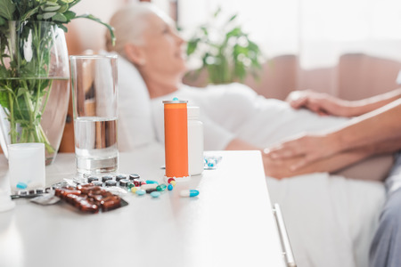 close-up view of pills and glass of water and senior patient lying in hospital bed behindの写真素材