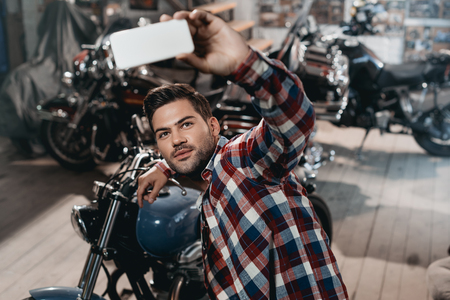 young handsome man taking selfie on smartphone with classic motorcycle in workshopの写真素材
