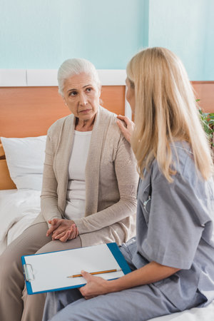 upset senior woman and nurse with clipboard looking at each other in hospitalの写真素材