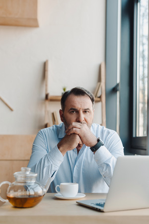 thoughtful mature businessman sitting in cafe with tea and laptopの写真素材