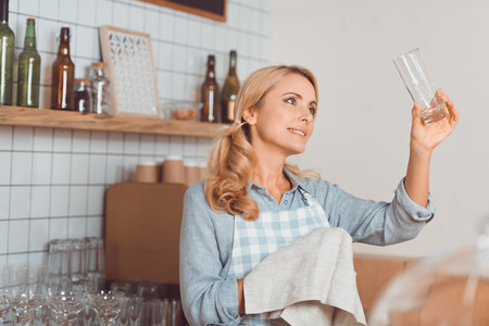 smiling waitress holding towel and looking at clean glass in cafeの写真素材