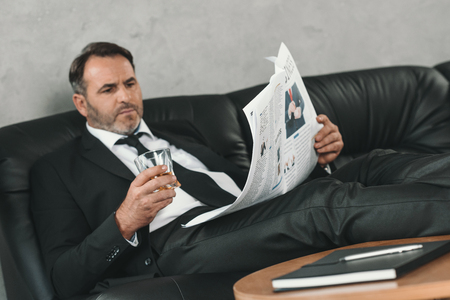 mature businessman with glass of whiskey reading newspaperの写真素材