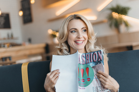 beautiful smiling mature woman reading fashion magazine and looking away in cafeの写真素材
