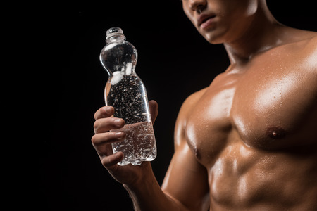cropped shot of muscular shirtless sportsman holding bottle of water isolated on blackの写真素材