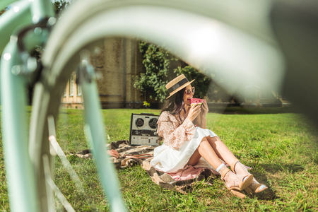 cheerful attractive girl eating watermelon while sitting on plaid in parkの写真素材