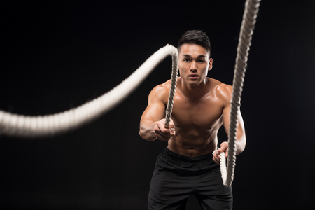 muscular shirtless asian man doing battle ropes exercise isolated on blackの写真素材