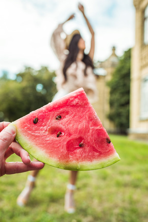 close-up partial view of hand holding slice of watermelon and young woman dancing behindの写真素材
