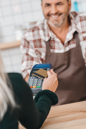 woman paying with pos terminal at cafe with smiling mature bartender waiting over counterの写真素材