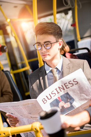 handsome young businessman in eyeglasses holding newspaper and looking away in busの写真素材