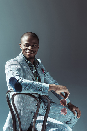 Smiling african american man in stylish clothes, sitting on wooden chair and holding sunglasses, isolated on greyの写真素材
