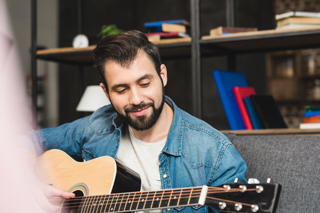smiling young man playing guitar while sitting on couchの写真素材