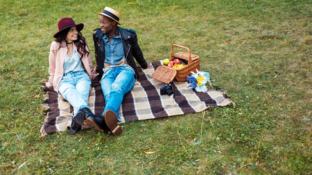 interracial smiling couple sitting on a blanket in the park and looking at each otherの写真素材