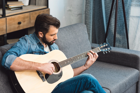 handsome young man playing guitar while sitting on couchの写真素材