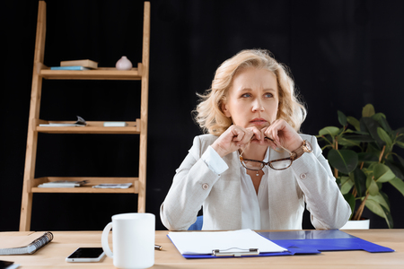 Pensive mature businesswoman holding eyeglasses and looking away while working in officeの写真素材