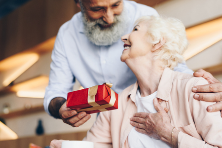 senior man surprising smiling wife with gift in cafeの写真素材