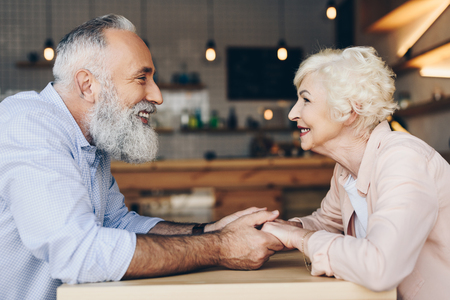 side view of smiling senior couple looking at each other and holding hands while sitting at table in cafeの写真素材
