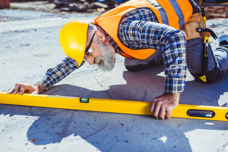 Bearded worker in reflective vest and hardhat working with spirit level at construction site,...