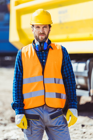 Worker in reflective vest and hardhat standing at construction site in front of big yellow truck and...
