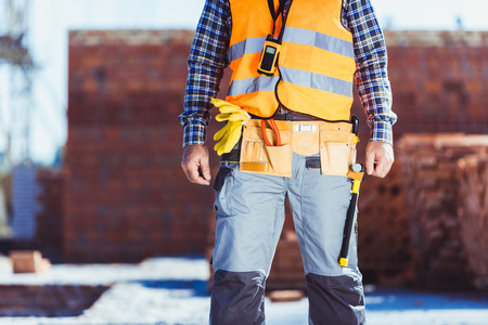 Cropped shot of builder in reflective vest and toolbelt standing at construction site