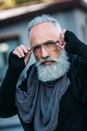 senior bearded man in fashionable clothing and eyeglasses posing on streetの写真素材
